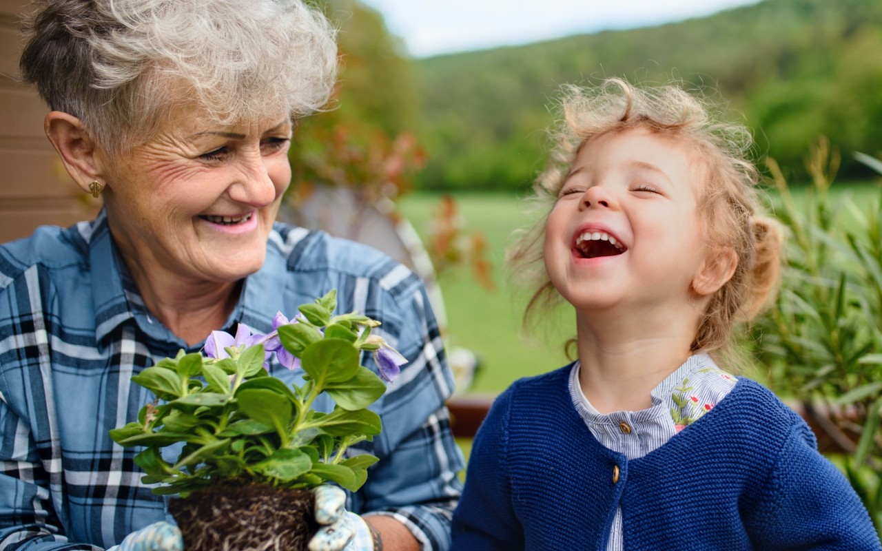 In a garden, a small child with curly hair laughs as her grandmother holds a leafy plant.