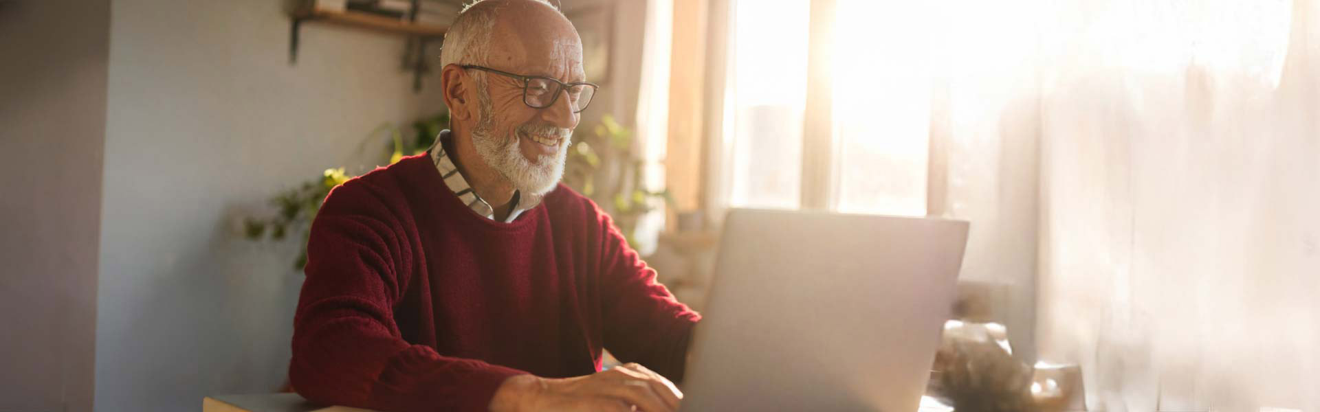 An older man wearing glasses is sitting at a desk resting his chin on his left hand while touching a laptop keyboard with his right.