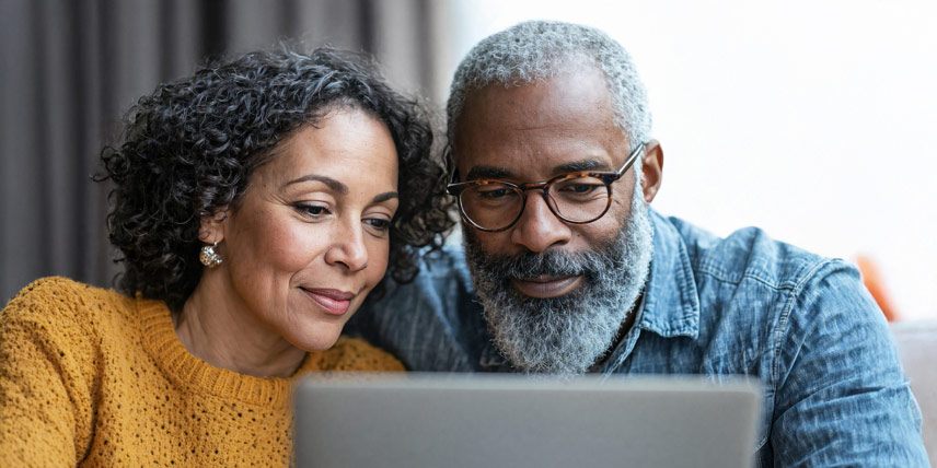 A husband and wife sit on a couch in front of a laptop.