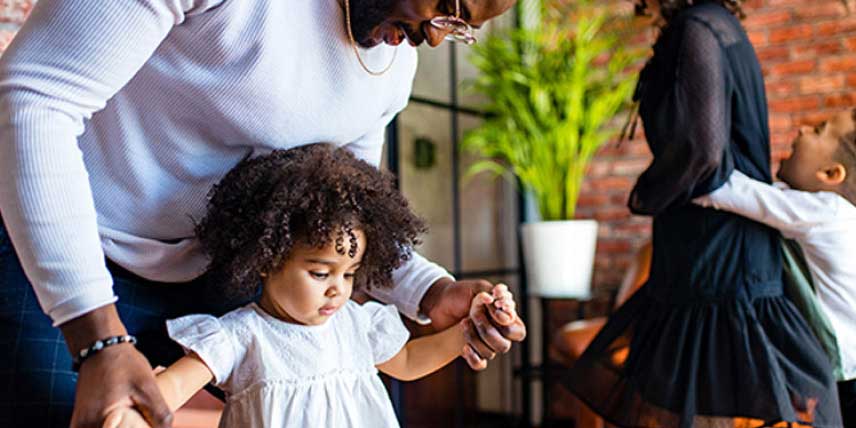 Father helps daughter walk while son plays in the background of a boutique.
