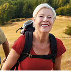 Person wearing a red shirt and backpack walking outdoors on a trail with grassy landscape and dense trees in the background.