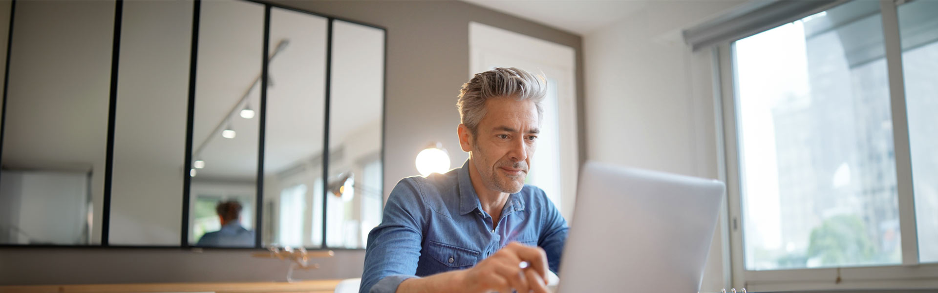 A middle-aged man is looking at his opened laptop screen while seated at a desk.