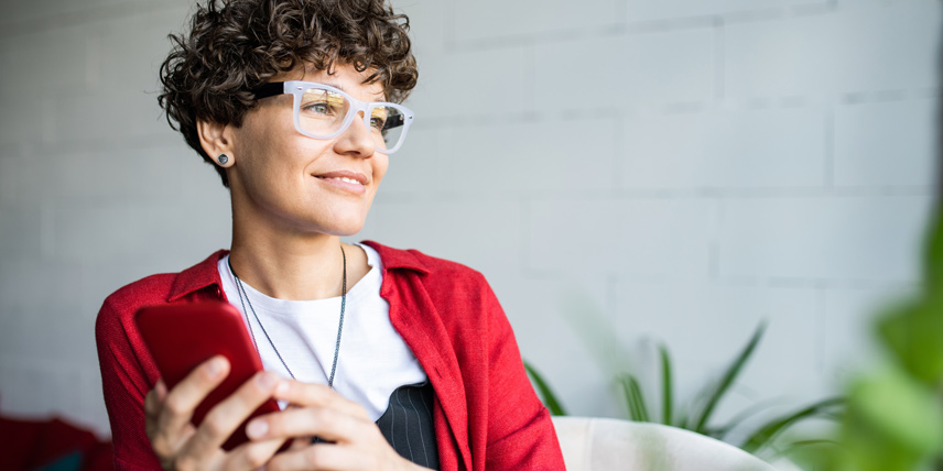 Woman with glasses holding a phone, smiling and looking into the distance.