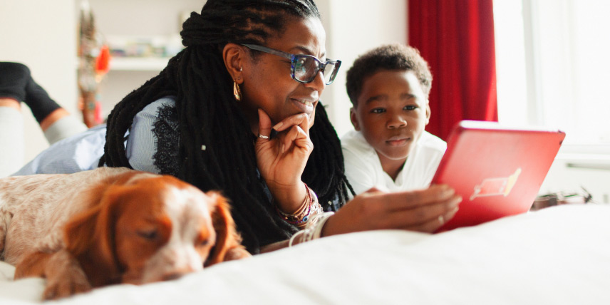 A mother lays beside her young son, smiling lovingly as they watch something on the tablet in her hand while the dog sleeps peacefully beside them.