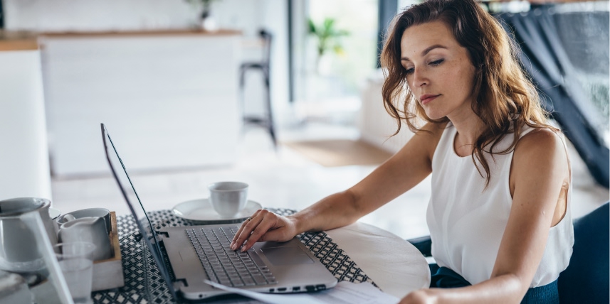Woman working on a laptop at home, reviewing papers at a kitchen table.