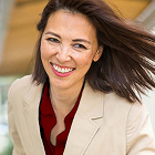 A woman wearing a beige blazer and red top smiles while standing outdoors, with her hair blowing slightly and a softly blurred background.