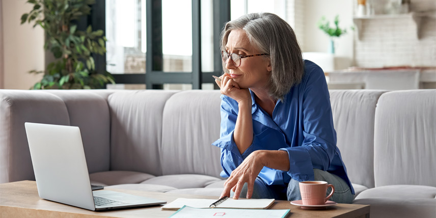 A woman with grey hair and a blue button-down sits on a couch in font of a notepad and laptop. She holds a pen, leaning forward to look at the laptop with satisfaction.