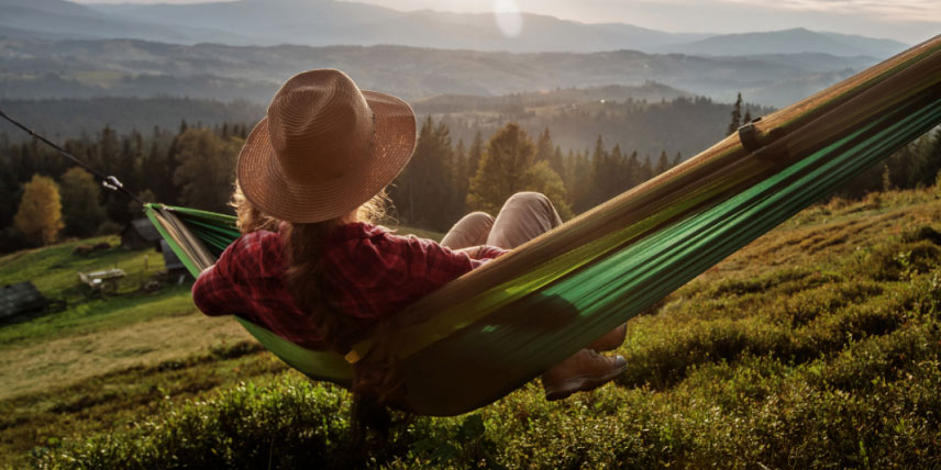 A person relaxes in a hammock on a sunny hillside overlooking a scenic landscape.