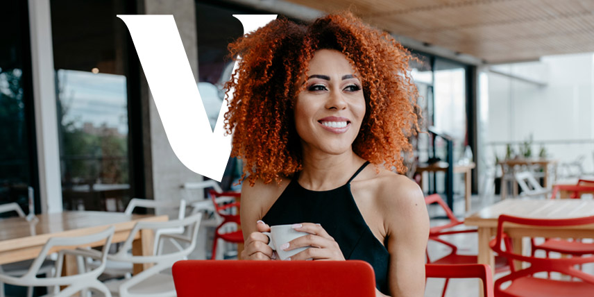 Woman seated at a laptop in an outdoor workspace, holding a mug and smiling.