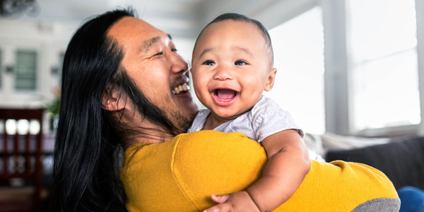 Photo of a man with long hair in a yellow shirt holding a young baby with a big smile.