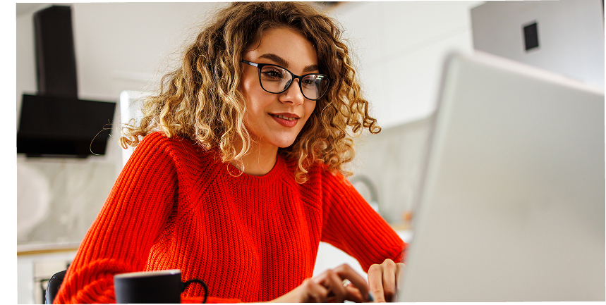 A young woman in a bright red sweater smiles confidently while typing on her laptop and having a cup of coffee at home.
