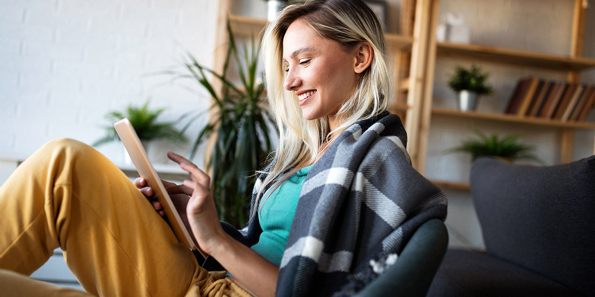 A woman relaxes in her home, smiling as she works on a handheld digital tablet.