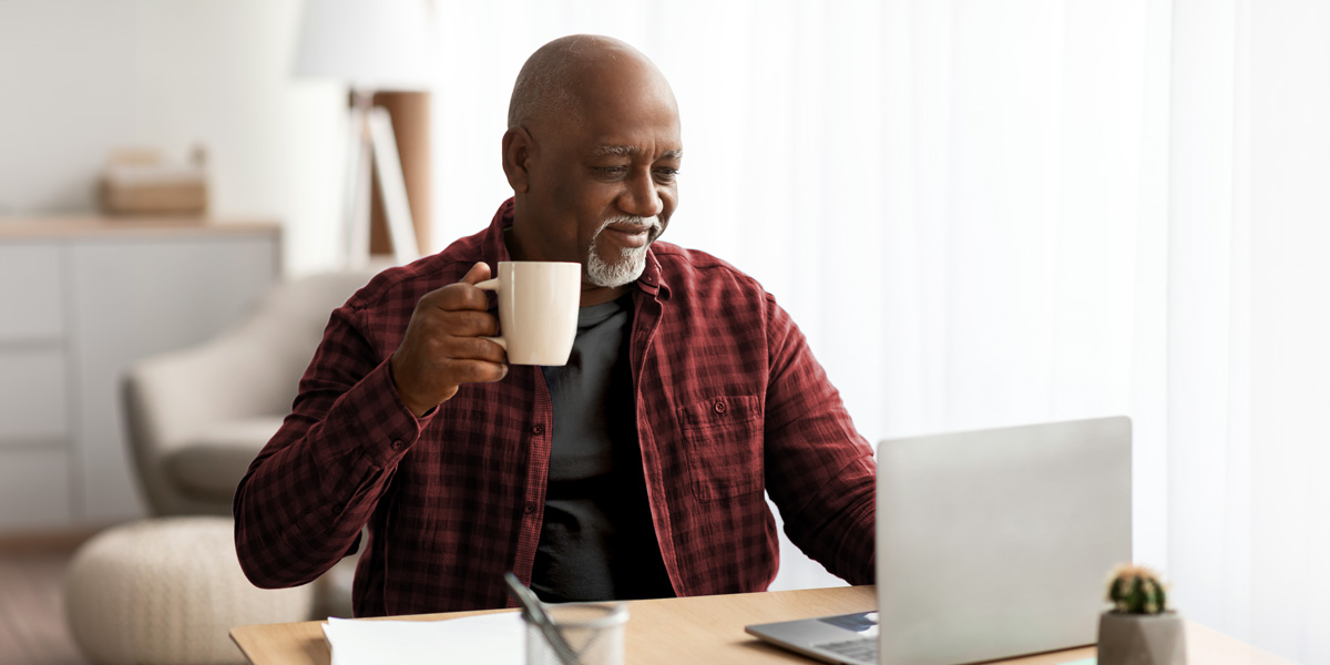 A man sitting at a table, looking at his laptop with a cup of coffee in hand.