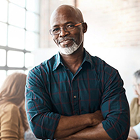 A man wearing glasses and a plaid shirt stands indoors with his arms crossed, in a softly lit office setting with people and desks in the background.