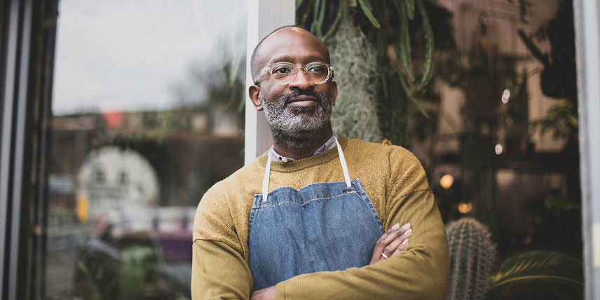 Man wearing an apron and standing outside a shop window with his arms crossed.