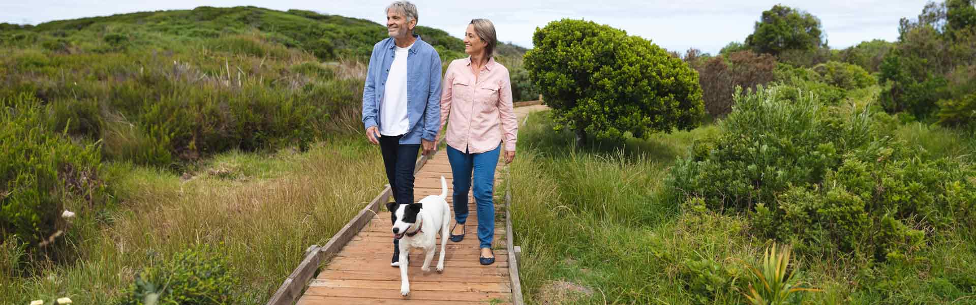 Retired age man and woman walk their dog on a path with greenery surrounding them.