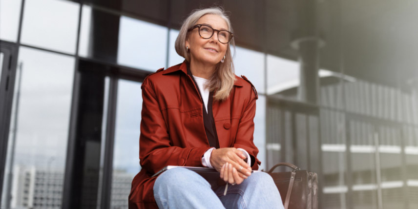 A woman in a red jacket stands on outdoor balcony, with a relaxed look on her face.