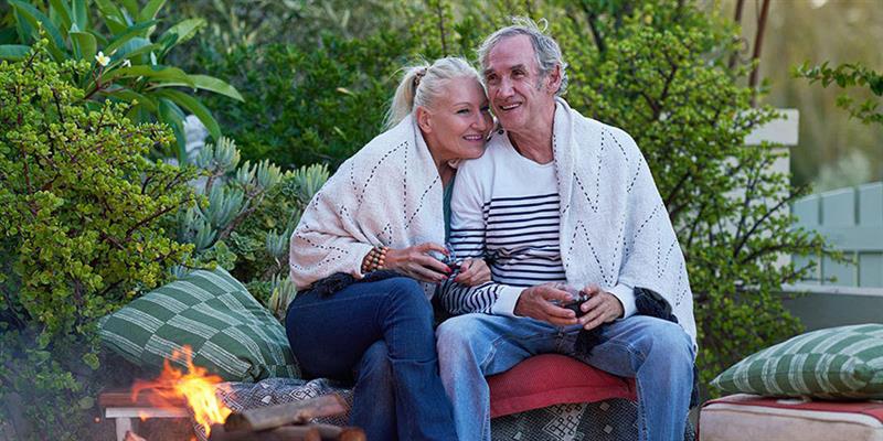 Mature, retirement age couple smile as they huddle together before a fire pit on an outdoor patio. 