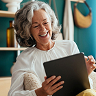 A woman with gray hair smiles while holding and looking at a tablet, seated comfortably indoors with shelves and household items in the background.