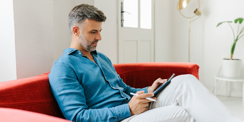 Man sitting on a red couch in a home, using a tablet and stylus.