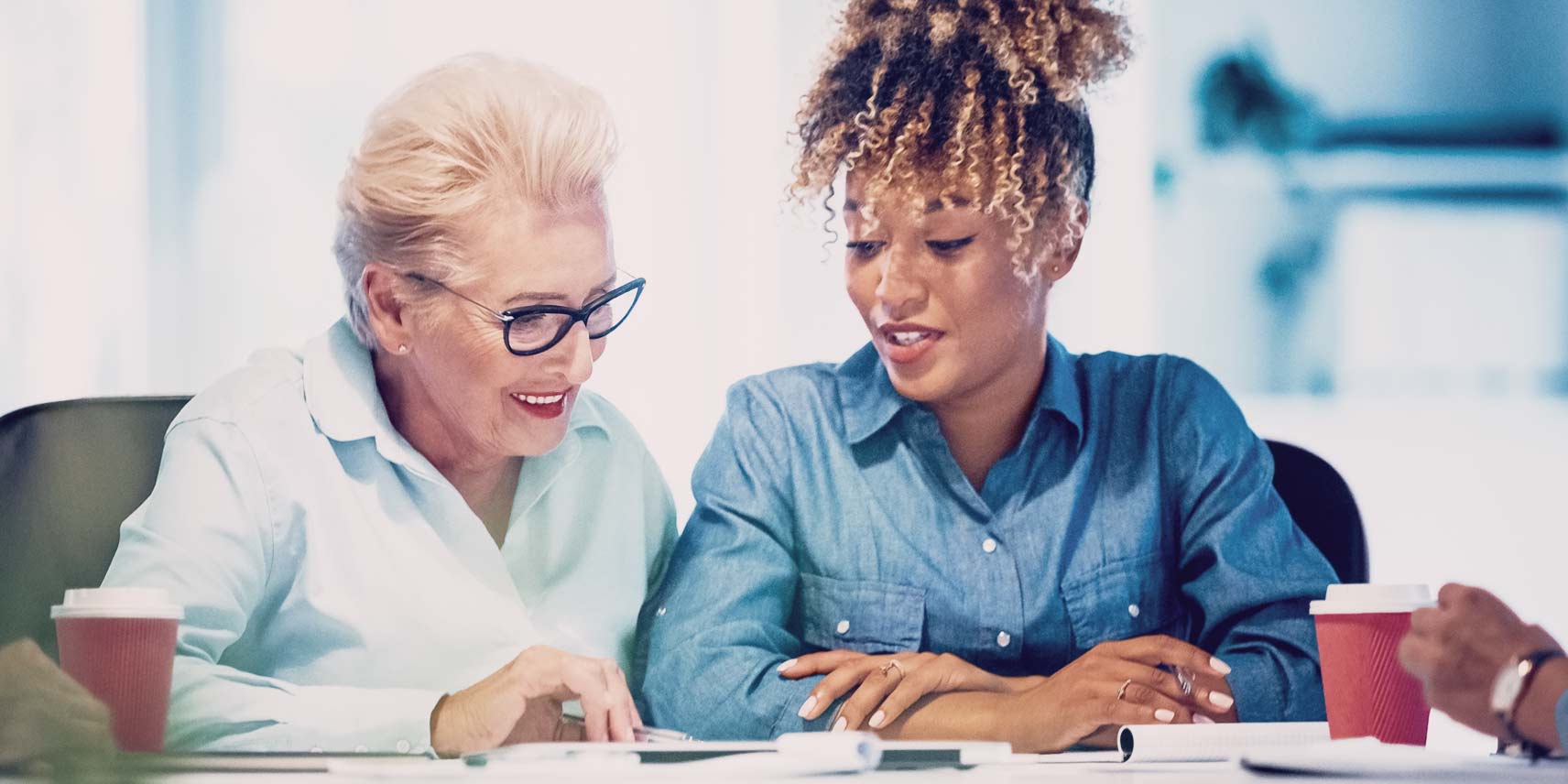 Two women are seated side by side at a table, engaged in conversation as they review documents together.