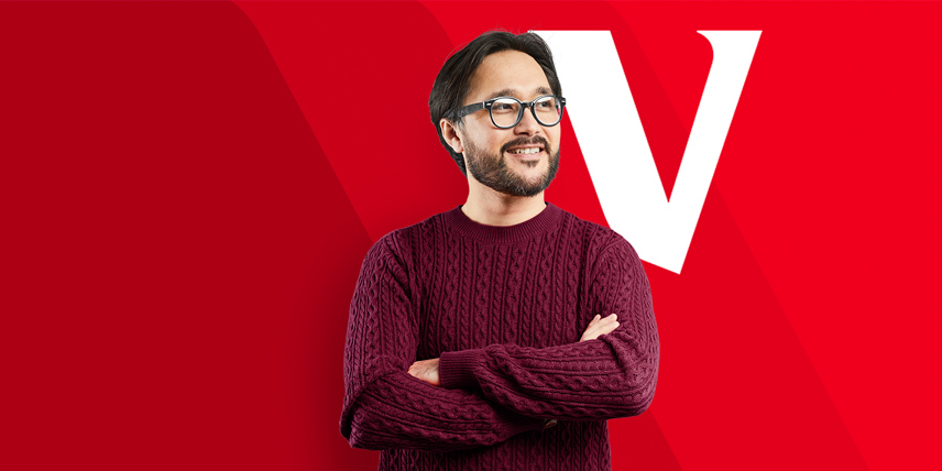 A man wearing glasses and a burgundy sweater stands with his arms crossed in front of a red background displaying the Vanguard V logo.