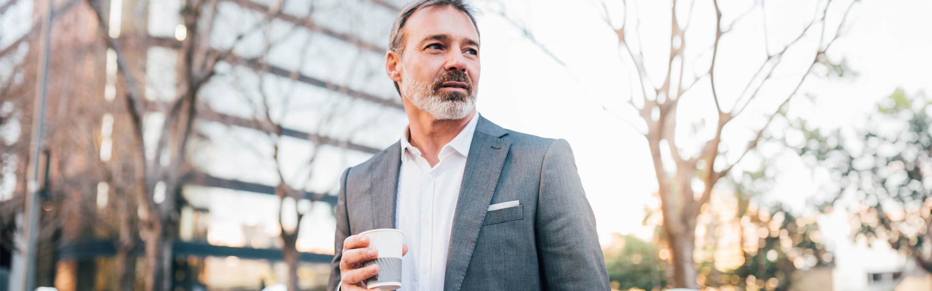 Man in a gray suit holds a cup of coffee in one hand, looks into the distance with an confident expression on his face.