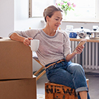A woman rests her arm on some cardboard boxes as she looks at her phone and enjoys a cup of coffee.