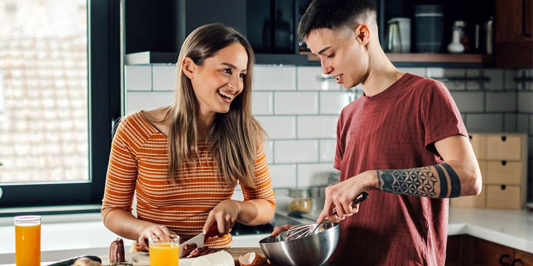Two people stand comfortably in a kitchen preparing food. One looks down, spreading something with a knife, while the other looks up at them, smiling brightly. The kitchen is sunny, clean, and lived-in.