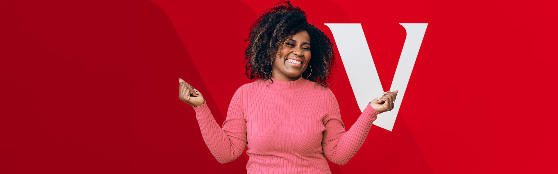 A woman with naturally curly hair smiles deeply as she stands against a red background with a large white letter 'V' partially visible behind her. She is wearing a pink sweater and is holding her hands up at her sides in excitement.
