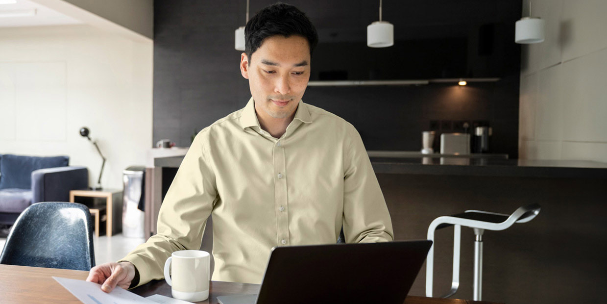 A man sitting at a desk, drinking coffee and looking at his laptop and some paperwork.