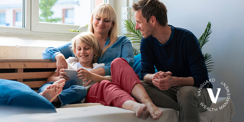 A mother and father relax with their child by a window, all of them smiling about something they’re watching on a tablet.