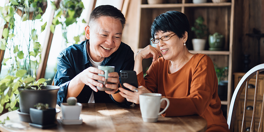 Asian couple looking at a smartphone while sitting at a wooden table with mugs and potted plants indoors