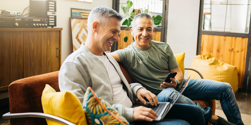 Two men sitting on a couch using a laptop and a smartphone at home.