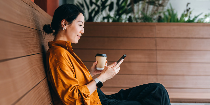 A woman sitting on a wooden bench, holding a smartphone in one hand a coffee cup in the other, appearing engaged with the phone.