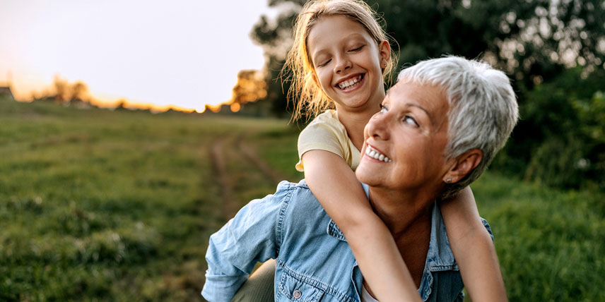 A woman is carrying a young girl piggy-back style through a green field, with the two of them smiling and laughing together.