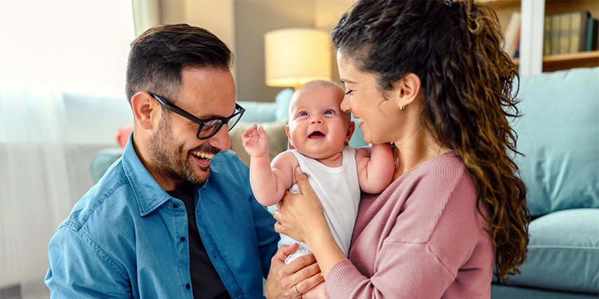Two smiling parents hold their infant in their living room.
