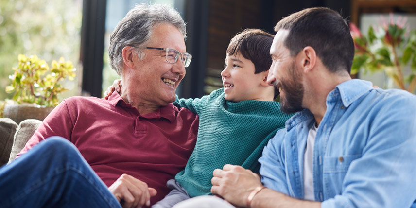 A grandfather, father, and son sit smiling on a couch.