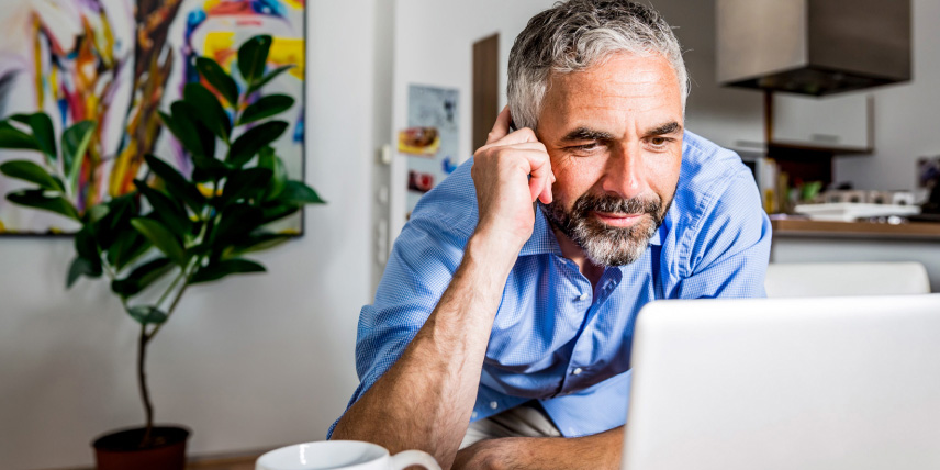 Man with beard using a laptop at a table in a home kitchen, with a coffee mug, plant, and artwork nearby.
