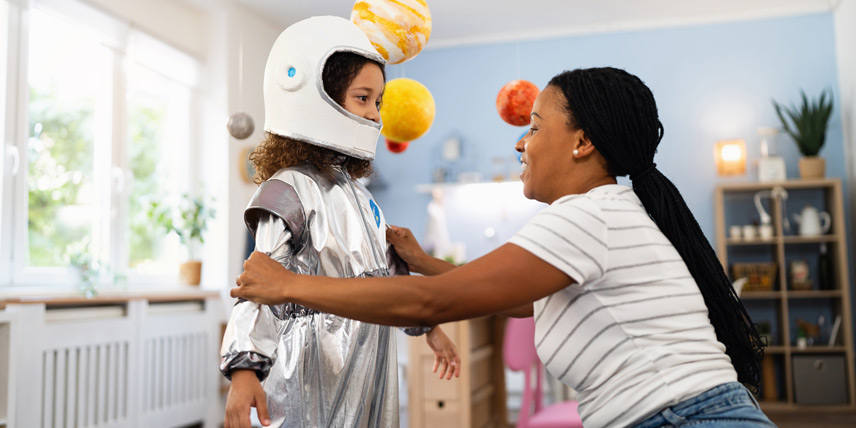 Child in an astronaut costume being helped with a space-themed dress-up activity in a bright playroom