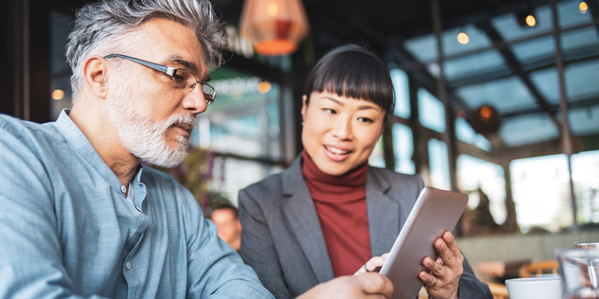 A man with glasses and a beard sitting beside a smiling woman in a blazer. They're looking at a tablet together.