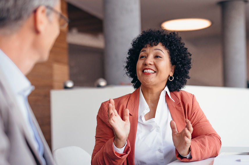 A smiling woman in a white shirt and red jacket gestures as she explains something to a man wearing glasses who is sitting across from her.