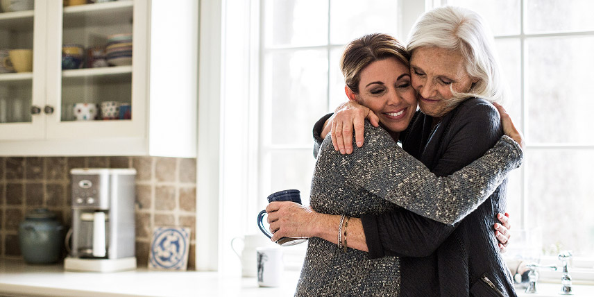 While holding a mug, a mother hugs her daughter in a bright kitchen.
