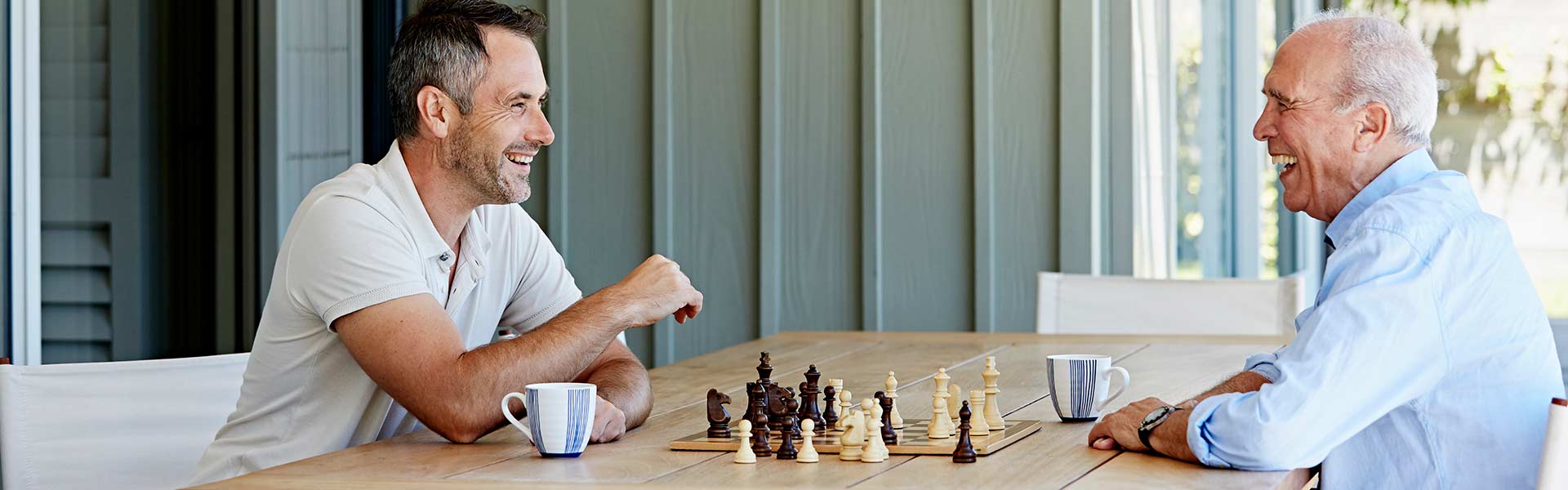 A man and his older father sit at a chessboard outside their home.