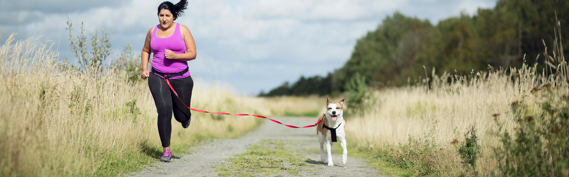 Woman running on a path with a dog on a red leash, surrounded by tall grass and trees in the background.
