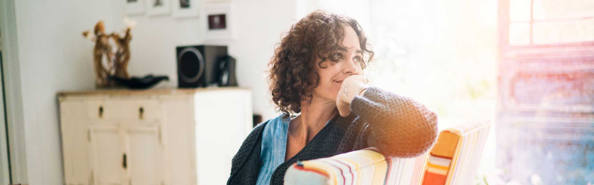 A pensive woman sitting on a colorful chair in her living room, with her hand on her chin..