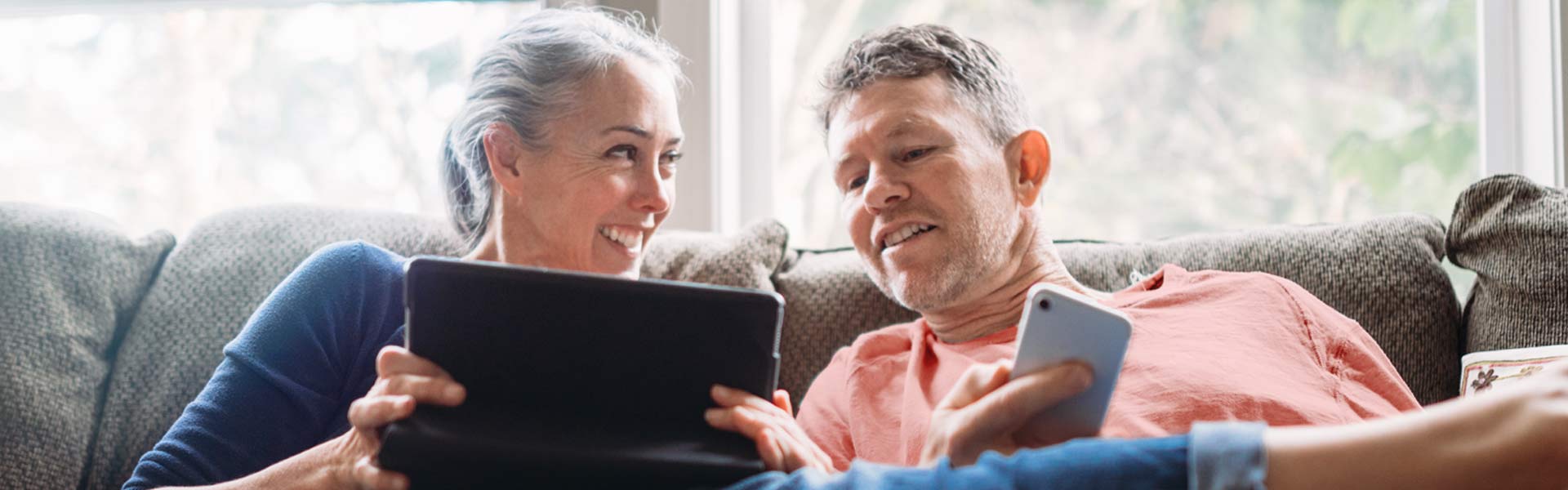 A man and woman are relaxing side by side on a sofa, looking at a mobile tablet the woman holds in her hands.
