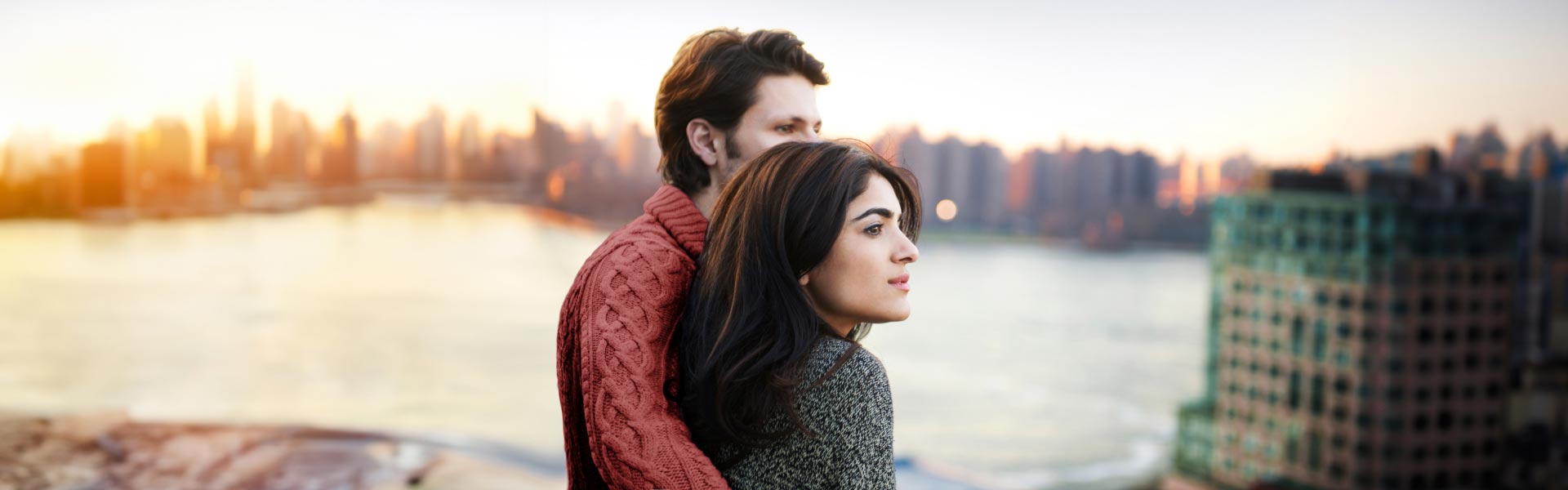 A man hugs a woman from behind as they look out together over the water toward the city skyline at sunset.