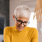 A woman with short hair and glasses looks down and smiles while wearing a yellow top, seated indoors with a softly blurred background.