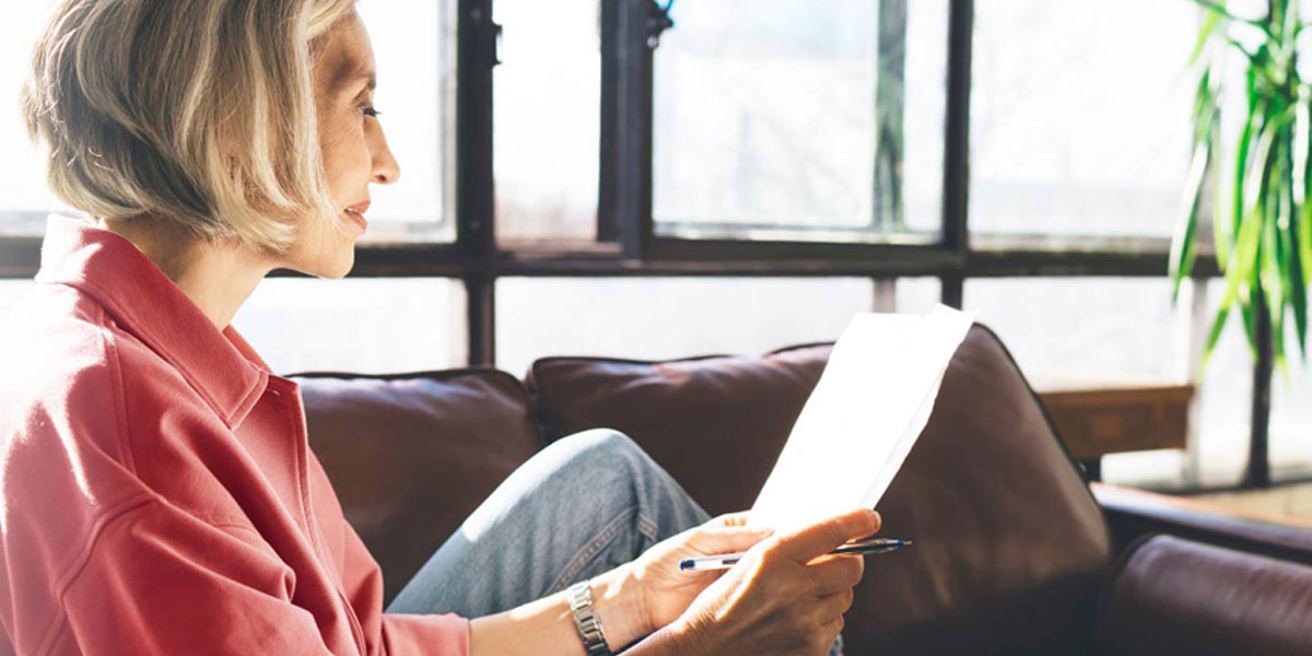 An older woman is sitting on a couch near a window looking at a document in her hands.